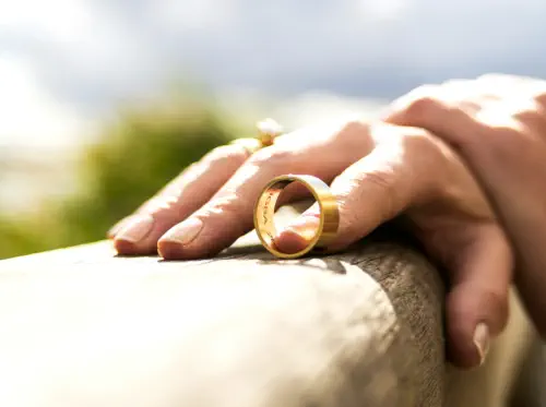 Closeup of a woman's hands. She has taken off her wedding band and is balancing it on a wooden fence rail with the pointer finger of her right hand.