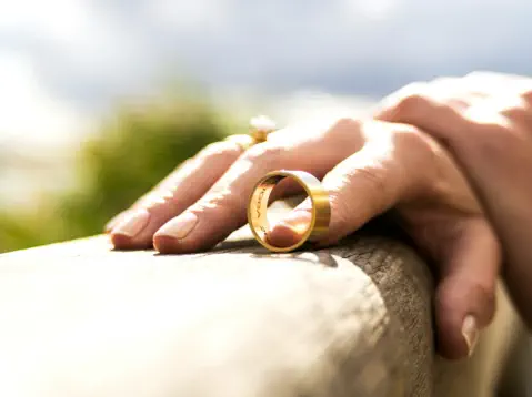 Closeup of a woman's hands. She has taken off her wedding band and is balancing it on a wooden fence rail with the pointer finger of her right hand.