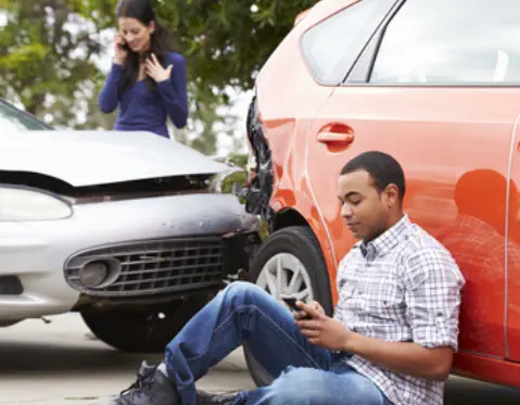 Two cars have been in a fender-bender. A woman stands, talking into a cell phone. A man sits on the ground looking at his cell phone.